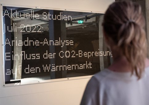 Eine jugendliche Person steht vor einem LED-Screen. Darauf steht "Aktuelle Studien, Juli 2022, Ariadne Analyse, Einfluss der CO2-Bespreisung auf den Wärmemarkt"