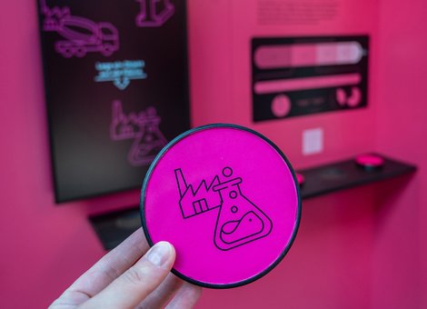 Blick in die Ausstellung. Eine Hand hält ein rundes, pinkes Objekt, auf dem zwei Icons zu sehen sind, die eine Industrieanlage und ein Versuchsglas aus der Chemie zeigen.