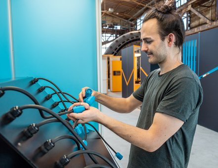 Ein Mann steht vor einer blauen Stele und versucht aus einer Menge unterschiedlicher Stecker die richtigen zu finden, die zueinander passen.
