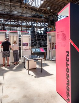 Blick auf die rote Themeninsel "Verwerten", an einer Stele mit Exponaten stehen zwei Besucher*innen.
