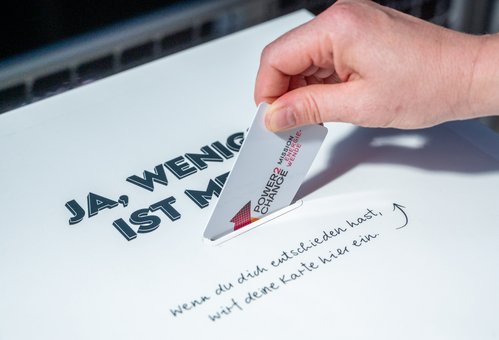 Eine Hand steckt eine Plastikkarte mit dem Power2Change-Logo in einen Wahlschlitz. Dieser ist mit "Ja, weniger ist mehr" beschriftet.