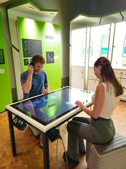 An einem Tisch mit interaktiver Touch-Funktion sitzen zwei Personen. Eine von ihnen hält einen Kopfhörer an sein Ohr.