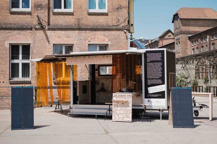 Foto des Energiemobils: Ein aufgeklappter Anhänger vor dem Solarpaneele stehen, im Inneren sind Ausstellungsstücke zu erkennen. Er steht auf einem Platz in einer Stadt.